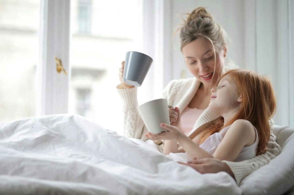 A mother and daughter sharing a warm moment with cups in bed, enjoying a cozy morning together.
